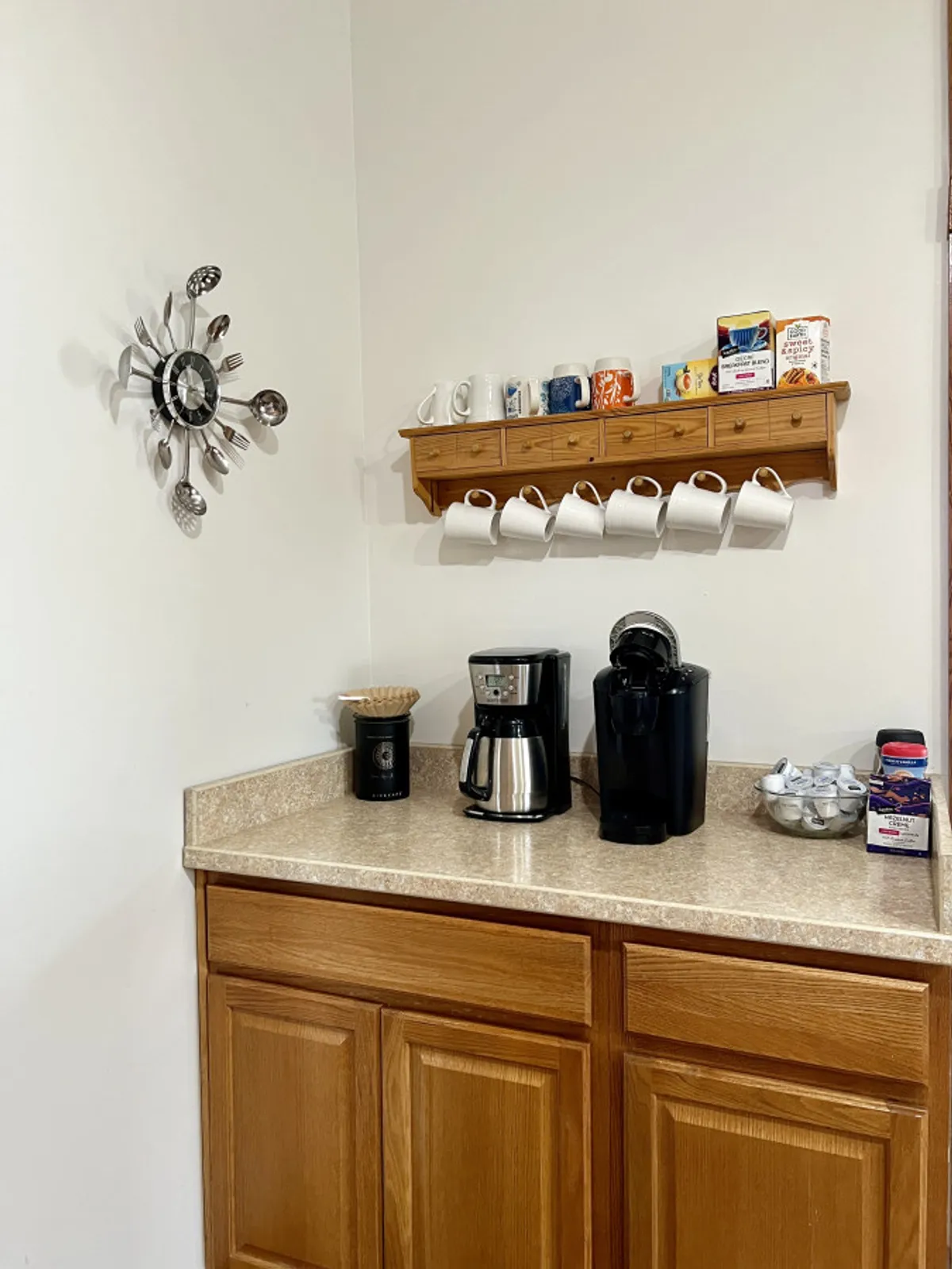 A close-up view of a coffee station featuring a coffee maker, electric kettle, a shelf with mugs, and decorative wall art, creating a cozy and inviting space for preparing morning coffee or tea.