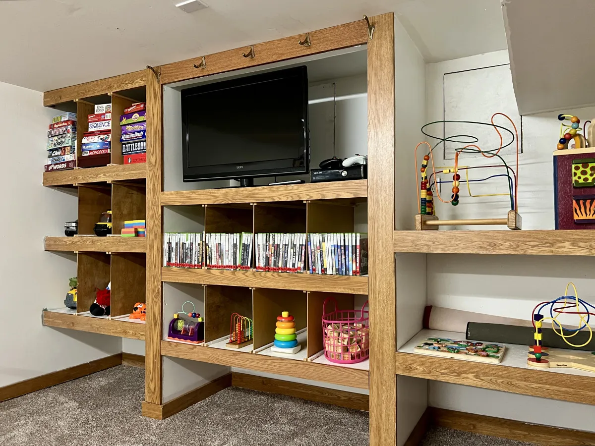 Shelves overflowing with colorful games and toys sit beside a TV in a bright play area, creating an inviting environment perfect for board games, TV time, and creative play. The mood is cheerful and energetic, ideal for families.
