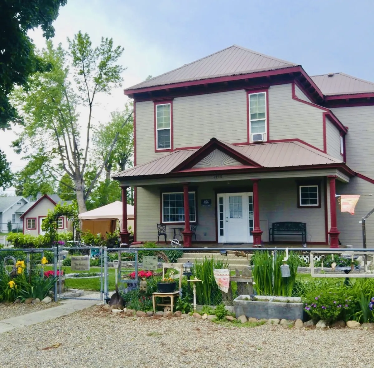 The image showcases the charming exterior of Scarlet Begonia, a beautifully renovated historic home set amidst vibrant flower gardens. The inviting front porch is furnished with comfortable seating, and the fenced yard provides a safe, picturesque space perfect for relaxing or socializing. The tranquil residential neighborhood and lush landscaping create a welcoming, serene atmosphere.