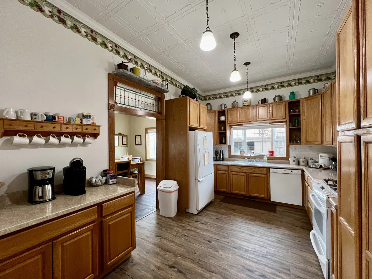 A warmly lit kitchen featuring a central island, a window allowing in natural light, hanging lights, a visible coffee maker, and a rack of cups. The inviting setup suggests a space designed for both culinary creativity and socializing, with enough room for multiple people to gather and cook or enjoy morning coffee together.