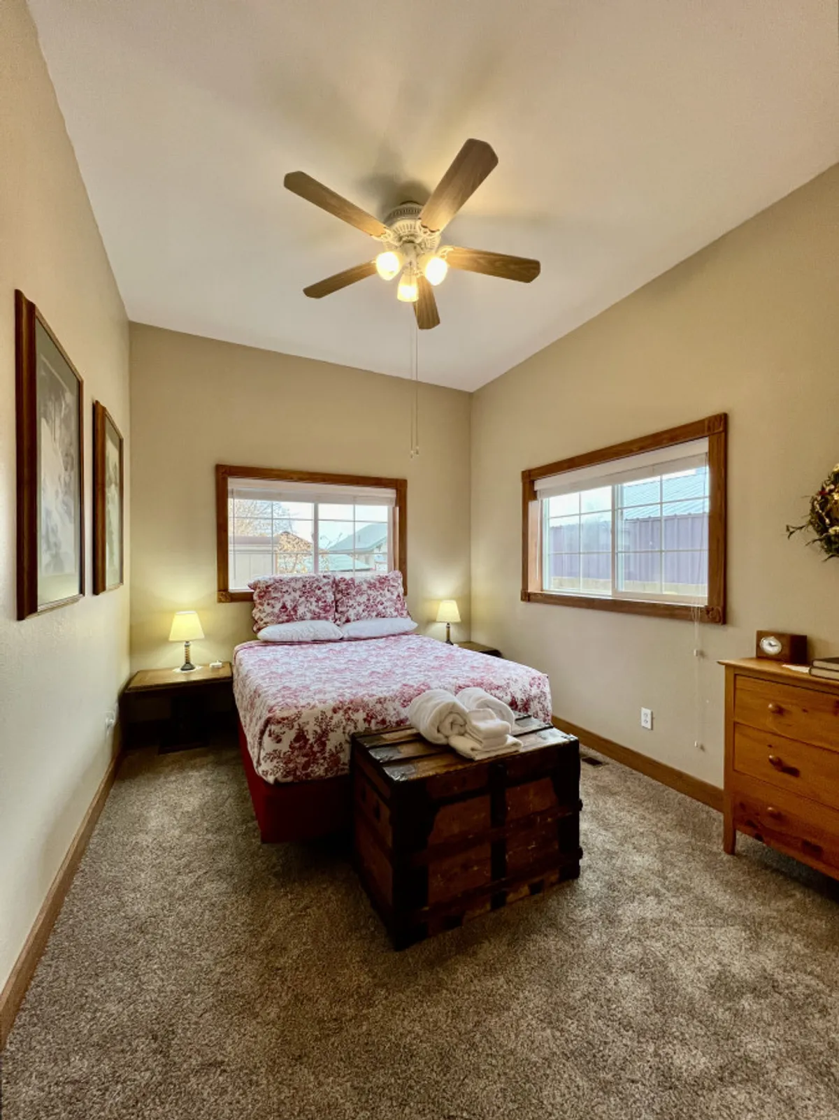 A warmly lit bedroom with a bed dressed in floral bedding, wooden furniture, and windows that flood the room with morning light. The atmosphere is both inviting and restful, ideal for relaxation or enjoying a quiet morning coffee.