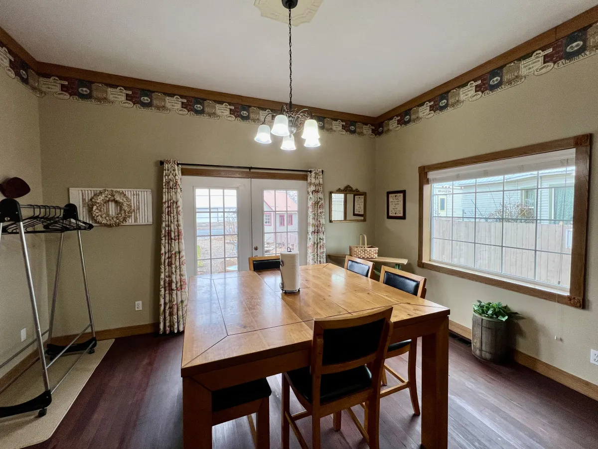 A wide view of the dining area with a large table, elegant chandeliers, French doors, and inviting window light. The scene radiates warmth and community, ideal for shared meals, celebrations, and group activities.