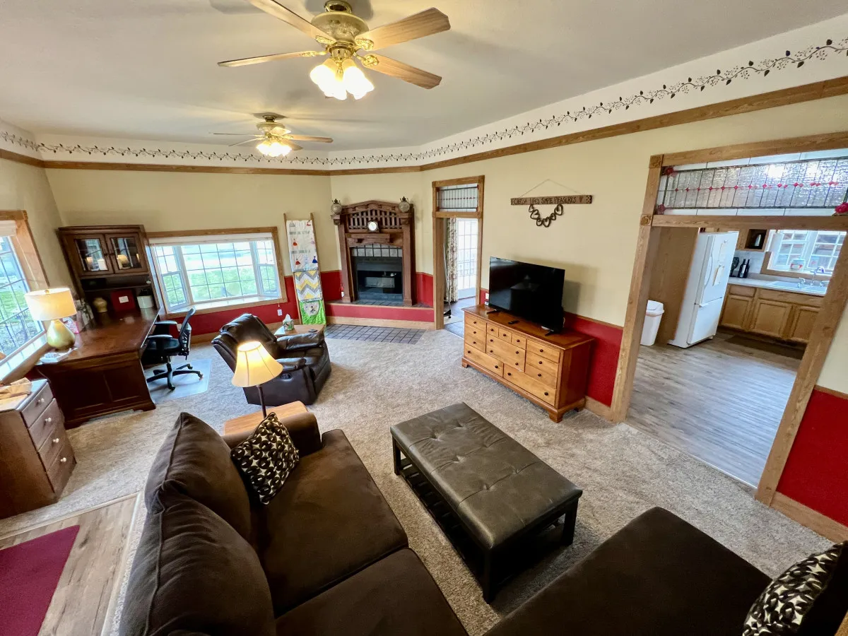 A warm and welcoming living room featuring a comfortable sofa adorned with decorative cushions, large windows that let in abundant natural light, an ornate fireplace mantel, classic wooden furniture, and a television situated for easy viewing. The space exudes a sense of togetherness and comfort, ideal for relaxing or gathering with loved ones.