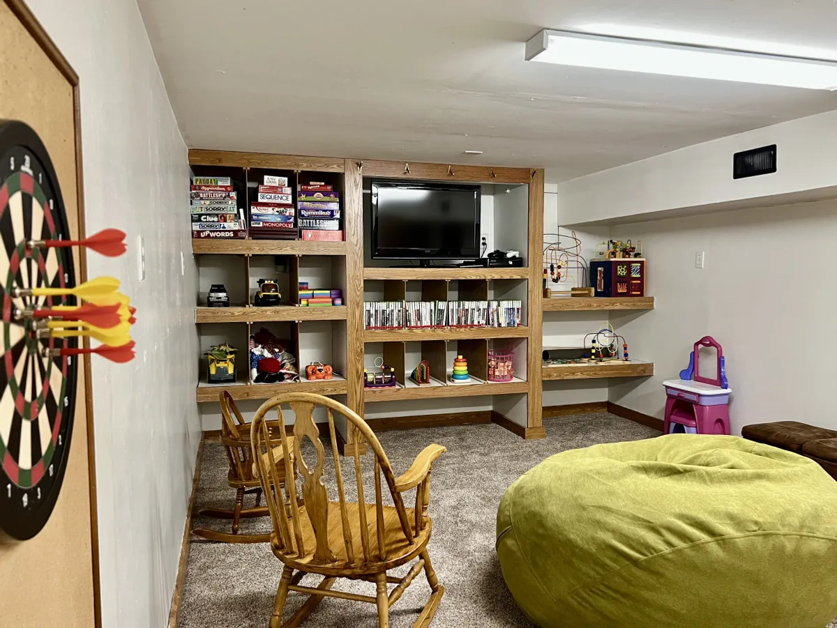 A spacious basement entertainment area featuring a dartboard, shelves filled with games and toys, a TV, and a comfortable seating area. The space is casual and family-friendly, ideal for gatherings, game nights, and movie marathons.